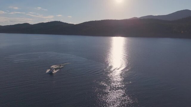 Backlit aerial view of boat pulling wakeboarder with bright sun reflected on lake