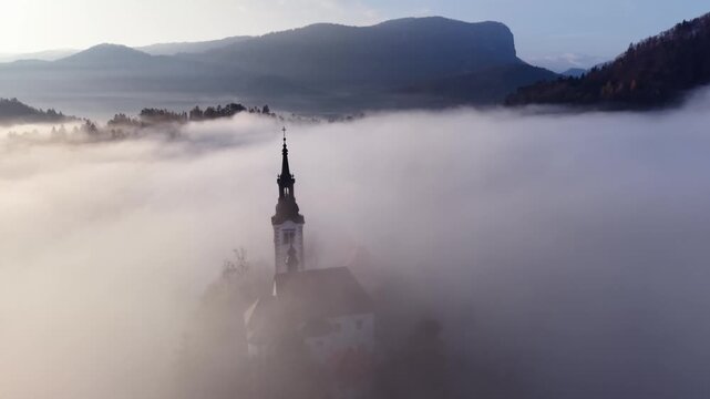 Church spire rising above thick blanket of fog at Lak Bled aerial view