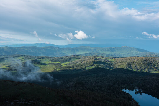 Scenic aerial top view above forest lake to green hilly vastness and layered mountain silhouettes far away under low dramatic cloudy sky. Wonderful scenery with sunlight and shadows of clouds on hills