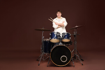 Smiling teenage boy with drumsticks and drum kit on brown background, space for text