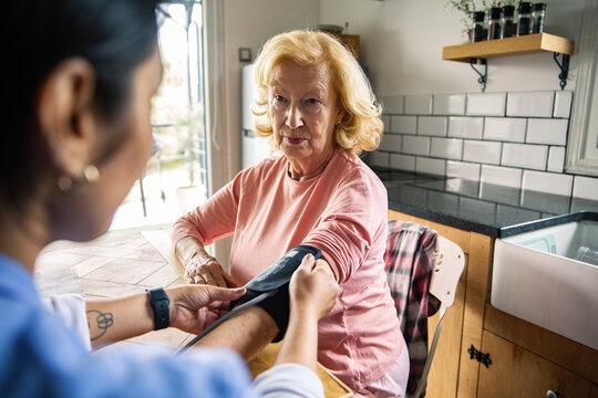 Home caregiver checking senior woman's blood pressure in kitchen