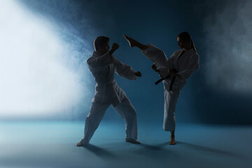 Man and woman in uniforms practicing karate on dark light blue background with smoke © New Africa