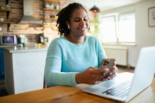 Smiling woman using smartphone and laptop at home kitchen