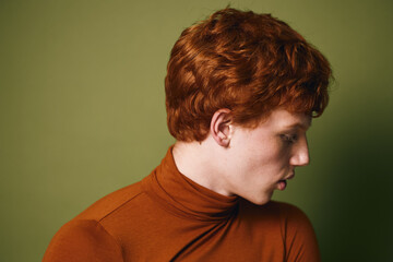 Young man with red curly hair wearing a brown turtleneck looking down against a plain olive background in studio lighting for portrait or fashion concept. © SHOTPRIME STUDIO