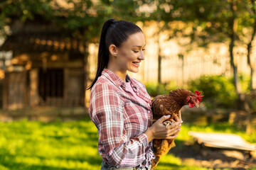 European female farmer in farm holding chicken, standing outdoors on backyard. Healthy lifestyle and organic farming concept © Home-stock