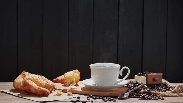 The slow-motion video shows a cup of hot coffee with steam rising, along with roasted coffee beans and bread on the table. This video was shot with a digital camera and edited by colleagues in a studi