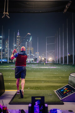Golfer in colorful attire swings club at Topgolf Dubai, illuminated city skyline visible in the background at night