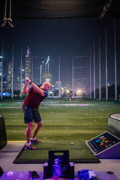 Man practicing golf swing at Topgolf Dubai with illuminated city skyline and golf balls scattered on the ground in the background