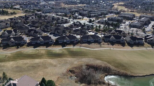 Grassy open space near South Fairplay Street features a curved walking path and aerated pond. Rippling water, manicured lawns, and uniform rooftops reflect soft midday light, Aurora, Colorado
