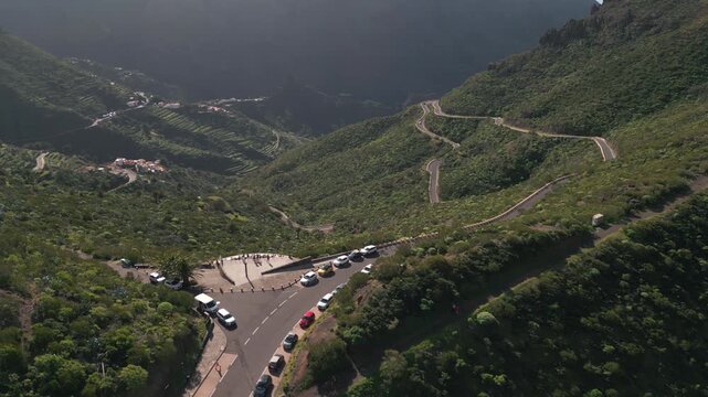 Winding mountain road with cars, lush green slopes, and a small village nestled in the valley.