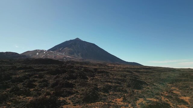 A majestic volcano rises from a barren, rocky landscape under a clear blue sky. Patches of snow dot the mountain's slopes.