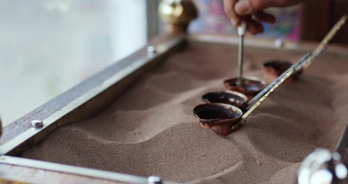 Close-up of a hand stirring Turkish coffee in small cezve pots placed in hot sand. Traditional Middle Eastern brewing technique showing an authentic artisanal coffee preparation process.