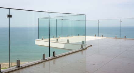 Obraz premium Modern luxury terrace with frameless glass railing and stone tile flooring overlooking a calm turquoise ocean under a clear sky.