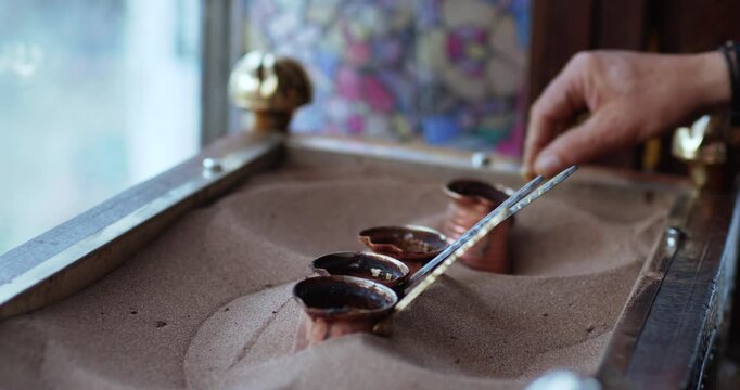 Hand preparing traditional Turkish coffee using small cezve pots heated in hot sand. Close-up view of an authentic Middle Eastern coffee brewing method and cultural ritual.