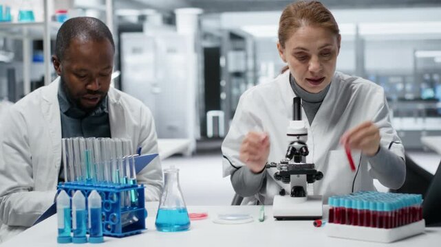 Pathologist examining biopsy slide while colleague documents diagnostic findings on clipboard. Medical experts working together to identify petri dish disease markers in clinical lab, camera B