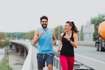 A young man and woman in athletic sportswear running side by side on a city bridge.