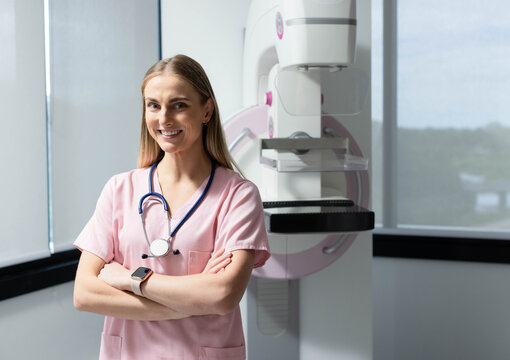Adult woman radiology tech standing in clinic near mammogram machine with pink scrubs, stethoscope