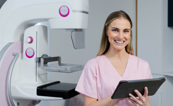 Female clinician in pink scrubs holding tablet beside mammography machine in clinical imaging room