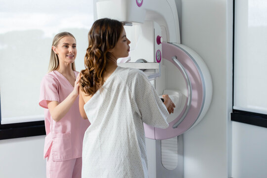 Female tech in pink scrubs guiding female patient in open-back gown in clinic onto mammogram paddle