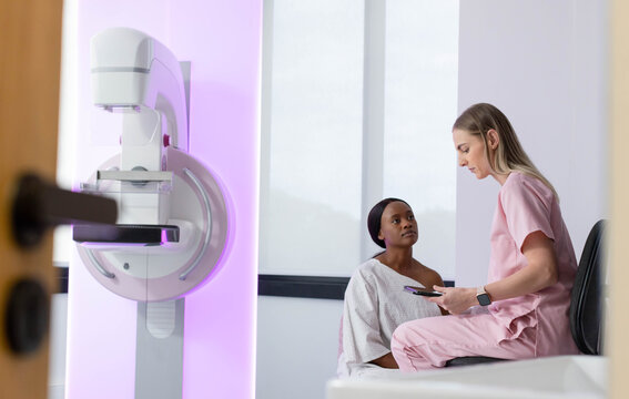 Female patient in hospital gown and female clinician consulting by mammogram unit, reviewing tablet