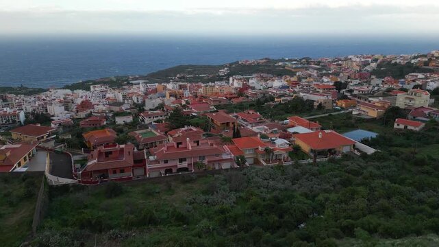Coastal town with red-tiled roofs nestled on a hillside, overlooking the blue ocean under a cloudy sky.