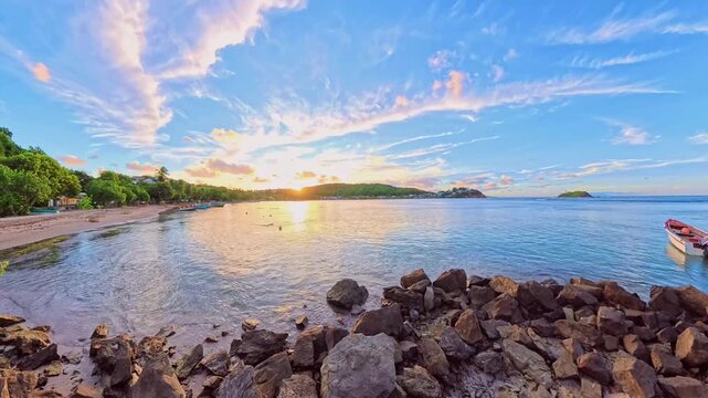 A panoramic time-lapse of the colorful sunset sky reflecting on Tartane beach of Martinique, with small fishing boats moored along the palm-fringed coast and rocky shore, French Antilles, Caribbean.