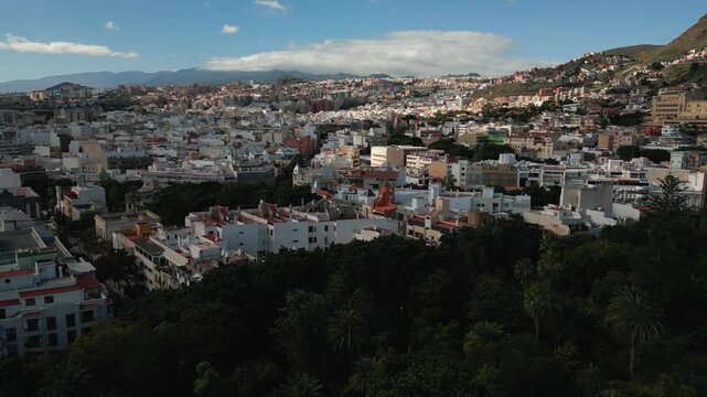 A sprawling city of white buildings climbs a lush green hillside under a bright blue sky with scattered clouds.