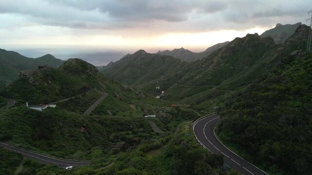 Winding roads cut through lush green mountains towards a hazy ocean horizon under a dramatic, cloudy sky.