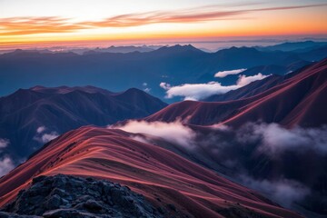 Sunrise over a layered mountain range reveals rolling peaks in warm hues Clouds drift between ridges silhouetted against a vibrant sky