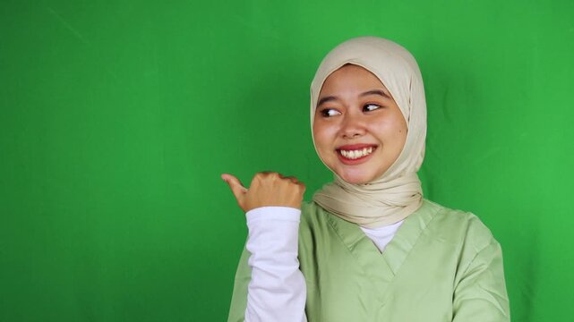 Cheerful Muslim nurse in white hijab and green scrubs pointing thumb left with bright smile looking sideways against green screen. Perfect for direction, recommendation, check this out, presentation, 