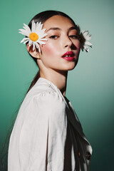 Obraz premium Portrait of a stylish woman with daisies in her hair wearing light clothes on green background. High fashion beauty concept with natural floral accessories and makeup.