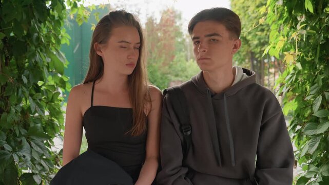 two teens seated on bench under leafy arch, girl in black dress and boy in grey hoodie share silent, tense gaze natural backlight filters through dense greenery, candid closeups capture pensive
