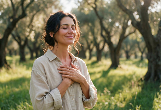 Smiling woman with closed eyes and hands on her chest in a sunlit olive grove, embodying gratitude, peace, and mindfulness