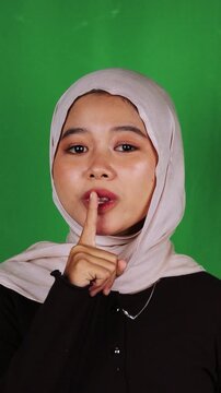 Muslim woman in beige hijab making shush gesture with finger on lips against green screen. Quiet signal perfect for silence, secret keeping, confidential, mystery, discretion, and library content.
