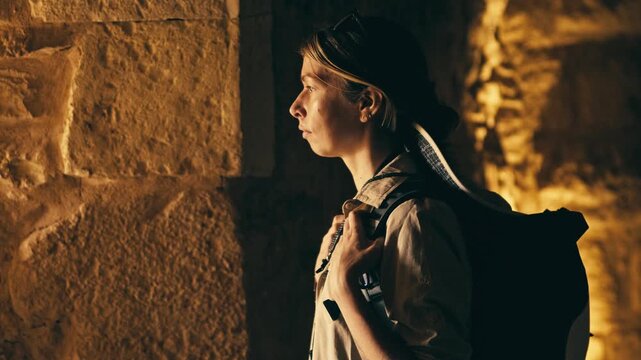 Cinematic side profile of a young woman explorer standing in an ancient stone passage, wearing a backpack and looking ahead. Warm golden light, moody atmosphere, travel and discovery concept