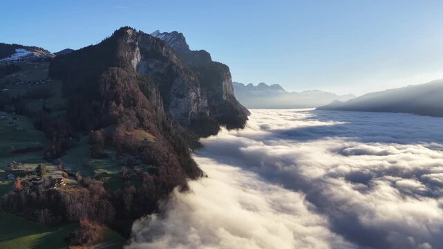 Aerial of Swiss houses on green slopes overlooking a vast sea of fog. Sharp mountain cliffs pierce the clouds under a bright sun in the Walensee region.