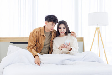 Fototapeta premium Asian young couple love happy sharing using mobile smartphone sitting lying on bed in white bedroom at home together boyfriend girlfriend relationship lifestyle relax beautiful romantic Valentine day.