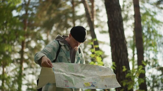 White hiker unfolding large map in forest, hands smoothing creases, focused gaze, scoutlike posture, sunlight filtering through canopy, planning route, map study, trail guide action