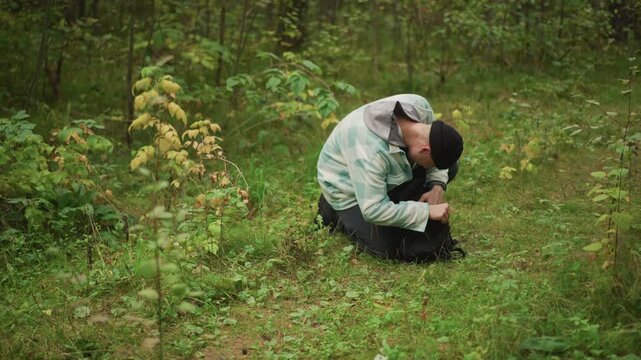 caucasian hiker inspecting ankle in forest, crouched on mossy ground beside backpack, checking lower leg for sprain or bite, focused expression, cool damp foliage, muted light, autumn greens,