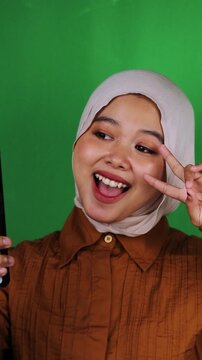 Playful Muslim woman in white hijab and brown shirt taking selfie with kissy face against green screen. Blowing kiss perfect for social media, selfie, flirty, fun, playful, romantic, affection content