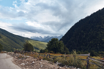 Obraz premium Landscape with a winding road, distant mountains, a rustic wooden fence, and a cloudy sky over a tranquil rural valley and meadows