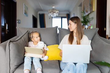 Mother working on laptop while daughter uses tablet on living room sofa