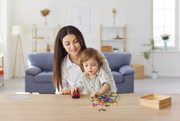 Naklejka premium Smiling young mother sitting at desk and playing with her little son holding toy car at home. Parent and kid making words of letters or learning alphabet during educational activity in living room.