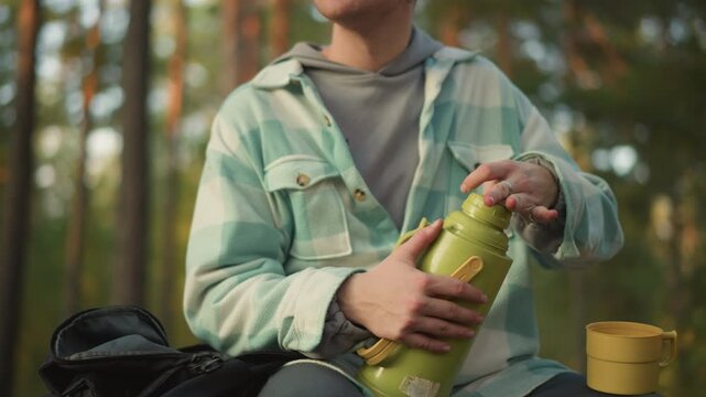 Traveler rests peacefully, Serene backpacker sips from thermos amid tranquil nature scenery, Relaxed traveler carefully tests lid of thermos before enjoying warm drink under sunsetlit pines