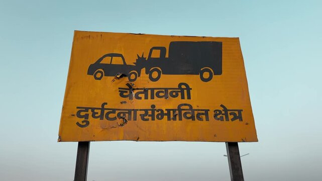 Camera orbiting shot of a yellow roadside warning board showing collision symbols and Hindi text, mounted on metal poles against open sky along a highway