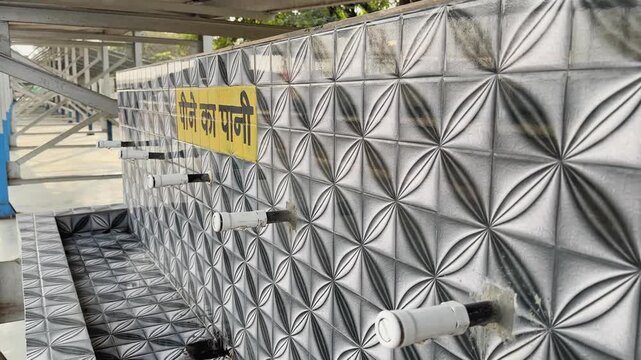 Wide view of drinking water area at railway platform with multiple taps fixed on patterned tiled wall, Hindi sign visible, clean public facility under covered station space