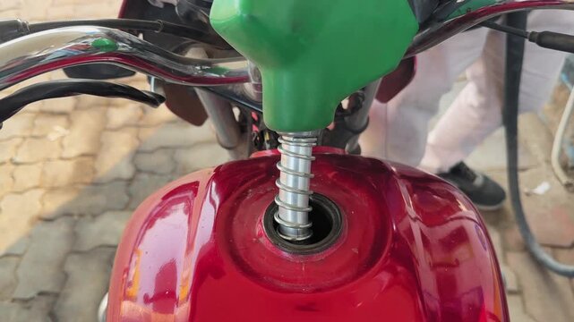 Closeup shot of a green fuel nozzle inserted into a red motorcycle fuel tank, showing petrol filling process with metal handle, reflections, and station pavement below