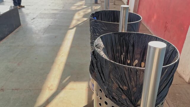 Camera pushing in shot onto stainless steel dustbins placed along railway platform walkway, showing waste collection setup with sunlight and shadows on station floor