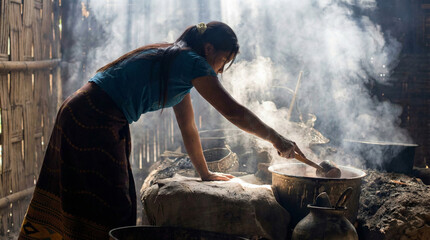 Asian woman cooking traditional meal in a rustic village kitchen with steam and natural light. Rural lifestyle and authentic culinary preparation in a traditional wooden kitchen setting.