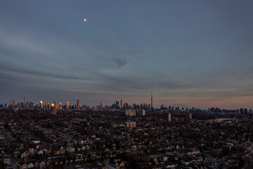 Naklejka premium Toronto cityscape featuring the iconic skyline and a residential neighborhood, with the moon visible in the twilight sky and buildings reflecting warm golden light during a serene evening